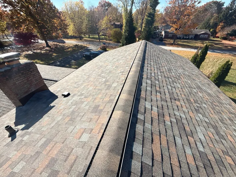 Straight ridge line on a freshly installed brown shingle roof with fall trees behind it